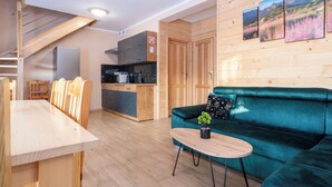 Una cocina moderna con armarios de madera, una zona de comedor con una mesa y sillas de madera, un sofá color esmeralda con una pequeña mesa, y un cuadro colgado en la pared que muestra un paisaje.