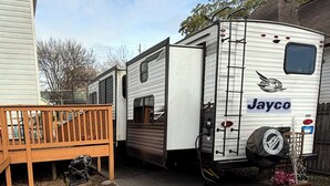 Een stacaravan met de merknaam 'Jayco' op de zijkant, geparkeerd in een woongebied met een houten terras en een fiets in de buurt.