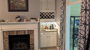 A living room with a fireplace, a blue sofa, a kitchen area with white cabinets, and a patterned curtain. - Cottage Beneath the Branches in Wachesaw-golf/pool/beach (Murrells Inlet)