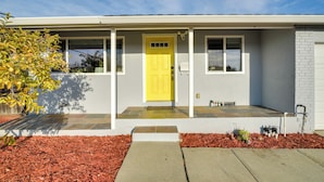 Une maison de plain-pied avec une porte d’entrée jaune, des murs extérieurs blancs et un petit jardin avant agrémenté de paillis rouge.