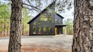 Una moderna casa a due piani con facciata scura, ampie finestre e vialetto in ghiaia circondato da alberi.