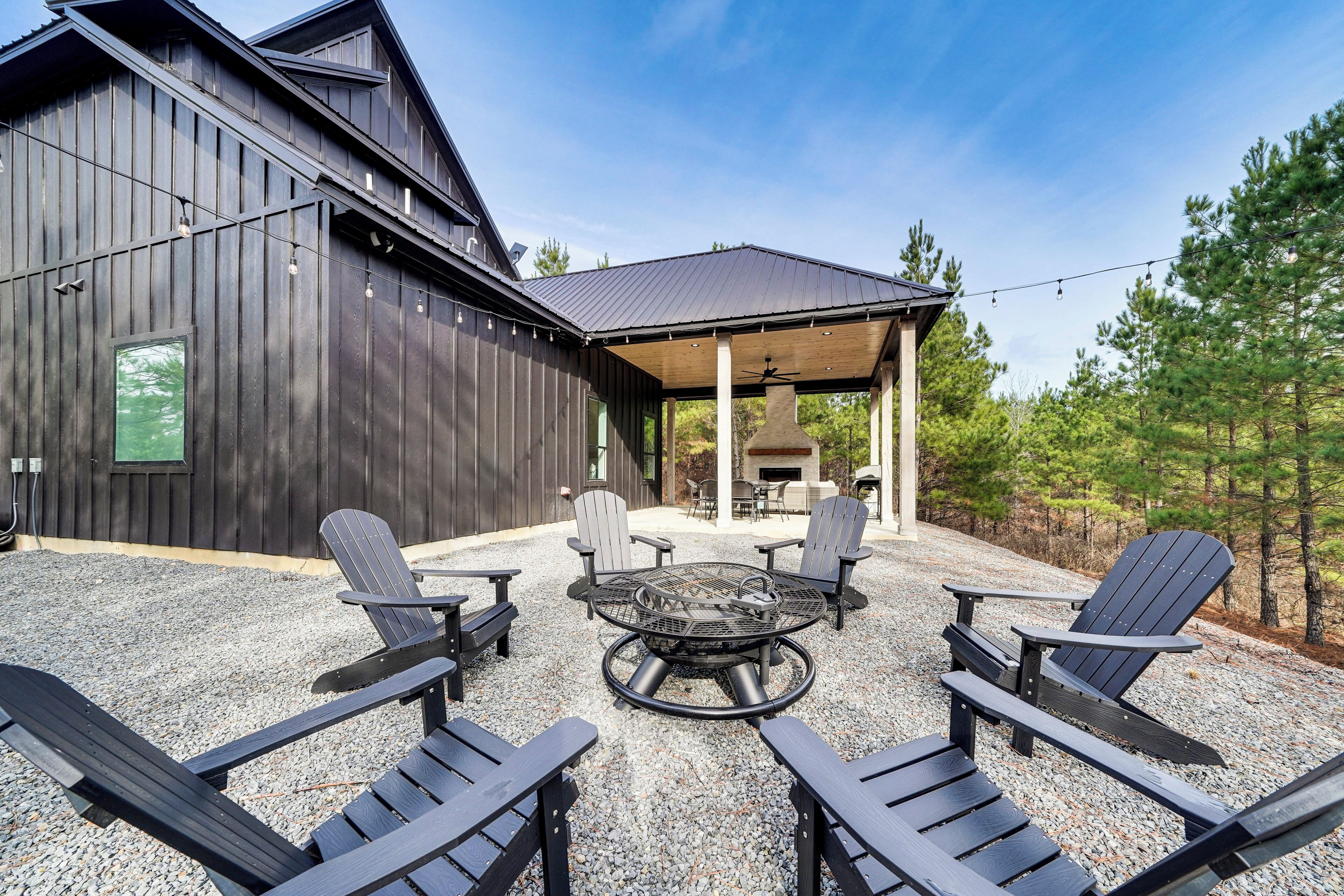 A covered outdoor seating area with Adirondack chairs and a fire pit, surrounded by a gravel patio and a wooden building.
