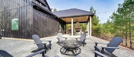 A covered outdoor seating area with Adirondack chairs and a fire pit, surrounded by a gravel patio and a wooden building.