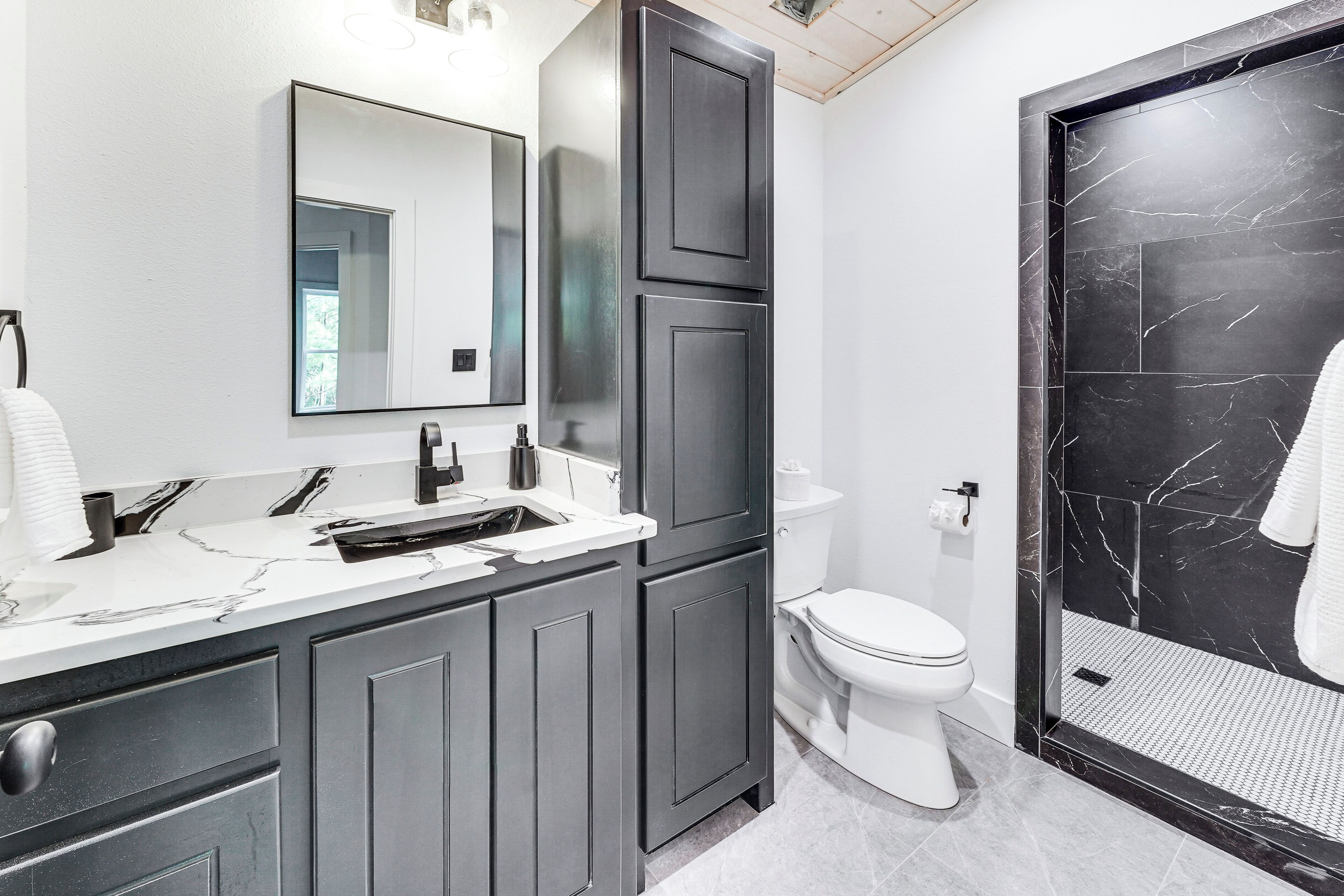 Een moderne badkamer met een grote spiegel, donkere kastjes, een wit toilet en een douchegedeelte van zwart marmer.