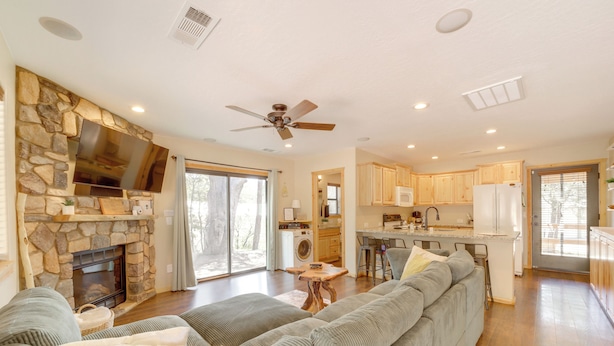 A spacious living room with a stone fireplace, a large flat-screen TV, a ceiling fan, and a kitchen area with wooden cabinets and a refrigerator. - Outdoor Entertainment Spaces: Remote Pine Home! (Pine)