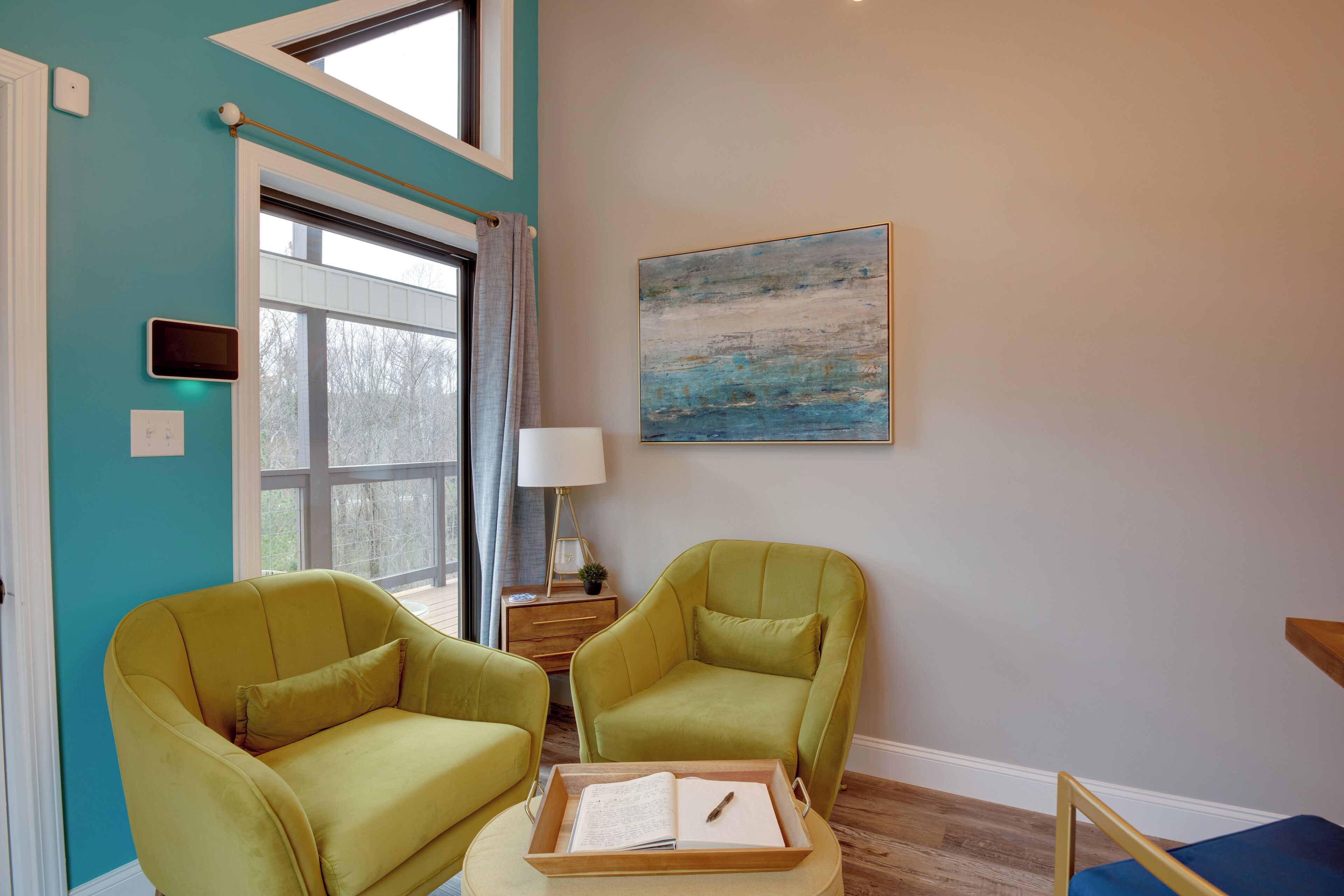 Une pièce aux murs de couleur turquoise, avec deux fauteuils verts, une table d’appoint en bois sur laquelle se trouve une lampe, un tableau abstrait encadré et une fenêtre avec des rideaux.