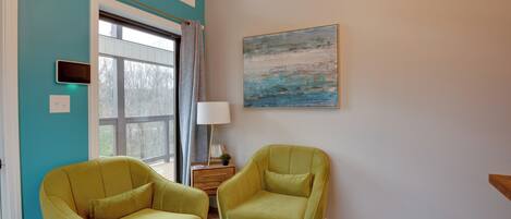 A room with teal walls, two green armchairs, a wooden side table with a lamp, a framed abstract painting, and a window with curtains.