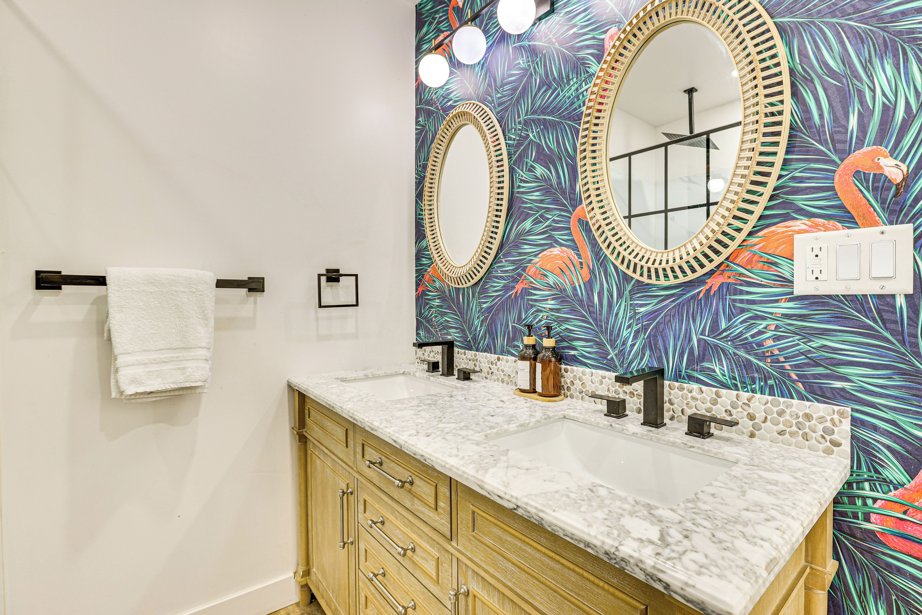 Une salle de bain avec deux lavabos, des meubles en bois, une serviette blanche et un miroir orné d’un motif de feuilles tropicales.