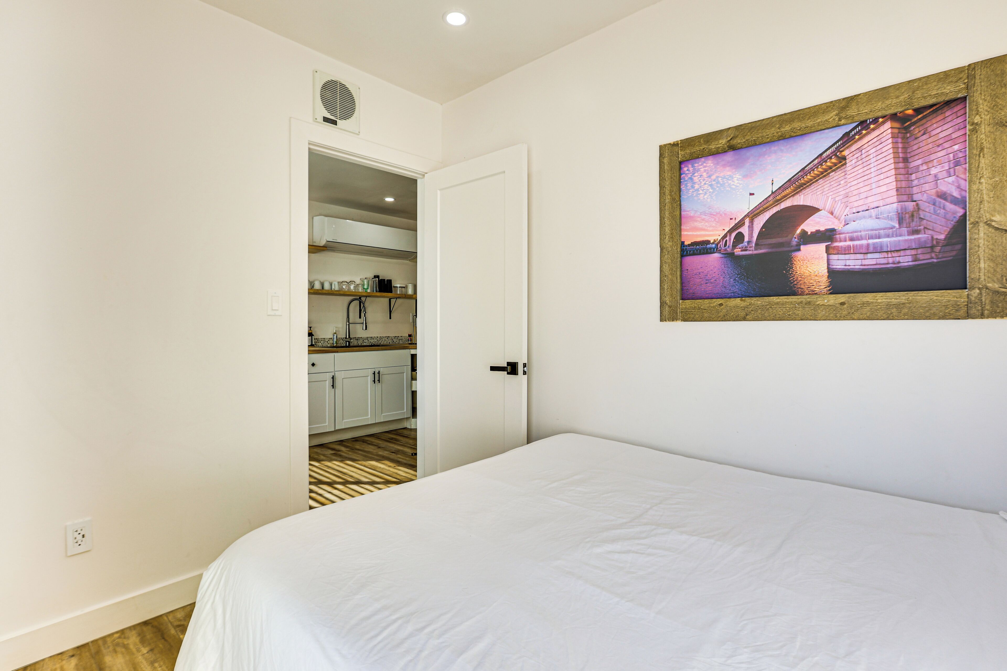 A bedroom with a bed, a door leading to a kitchen, and a framed picture of a bridge on the wall.