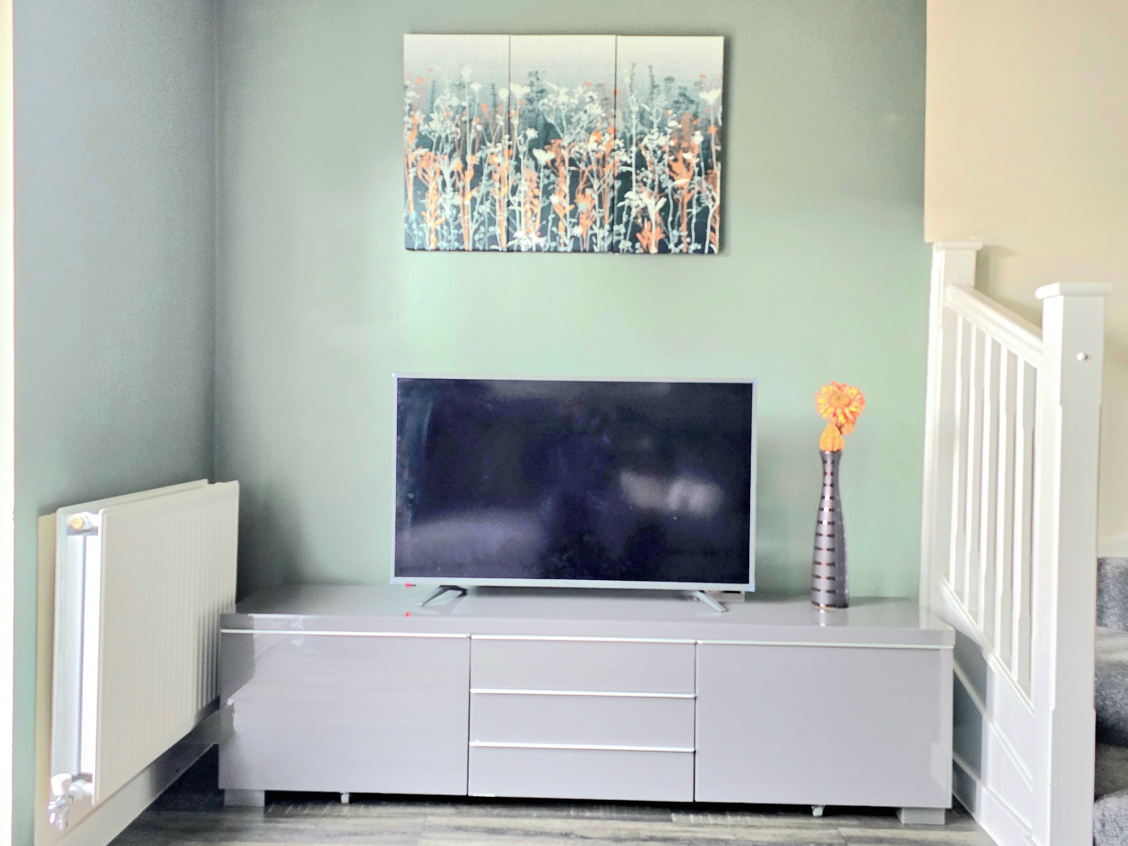 A living room with a flat-screen TV on a white cabinet, a painting above the TV, and a vase with flowers on the cabinet.