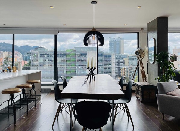 A modern dining area with a large table and chairs, a hanging light fixture, and a view of the cityscape through large windows.