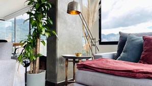 Ein modernes Wohnzimmer mit einem grauen Sofa, einer großen Topfpflanze, einer Stehlampe und Blick auf die Stadt durch große Fenster.