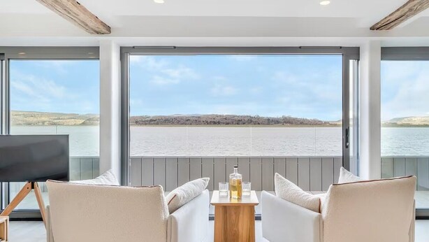 A modern room with two white sofas facing a large window overlooking a body of water. - Beach Haven - Stunning Beach Front Property (Arnside)