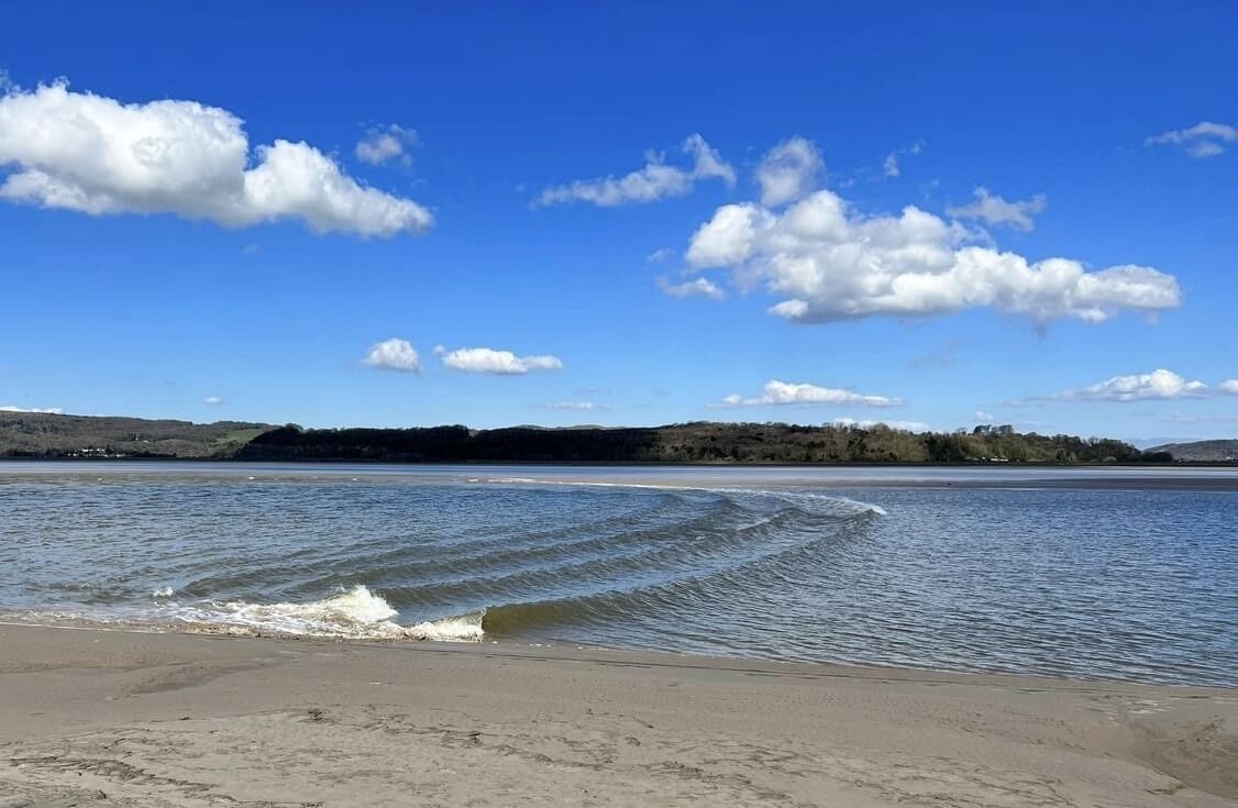 Une plage avec une eau calme, un ciel dégagé et des collines qui se dessinent à l’horizon.
