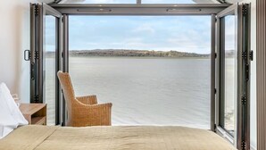 A bedroom with a large window overlooking a body of water, a wicker chair, and a bed. - Beach Haven - Stunning Beach Front Property (Arnside)