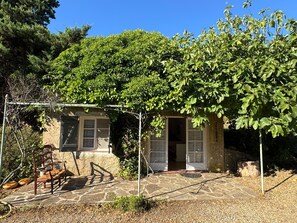 Un bâtiment rustique en pierre, doté d’un patio couvert, d’une chaise en bois et d’une pergola ornée de plantes grimpantes.