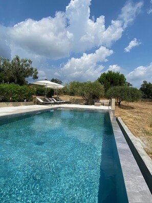 Une piscine rectangulaire à l’eau d’un bleu limpide, entourée d’un bord en béton peu élevé. On y trouve des chaises longues et un parasol blanc sous un arbre.