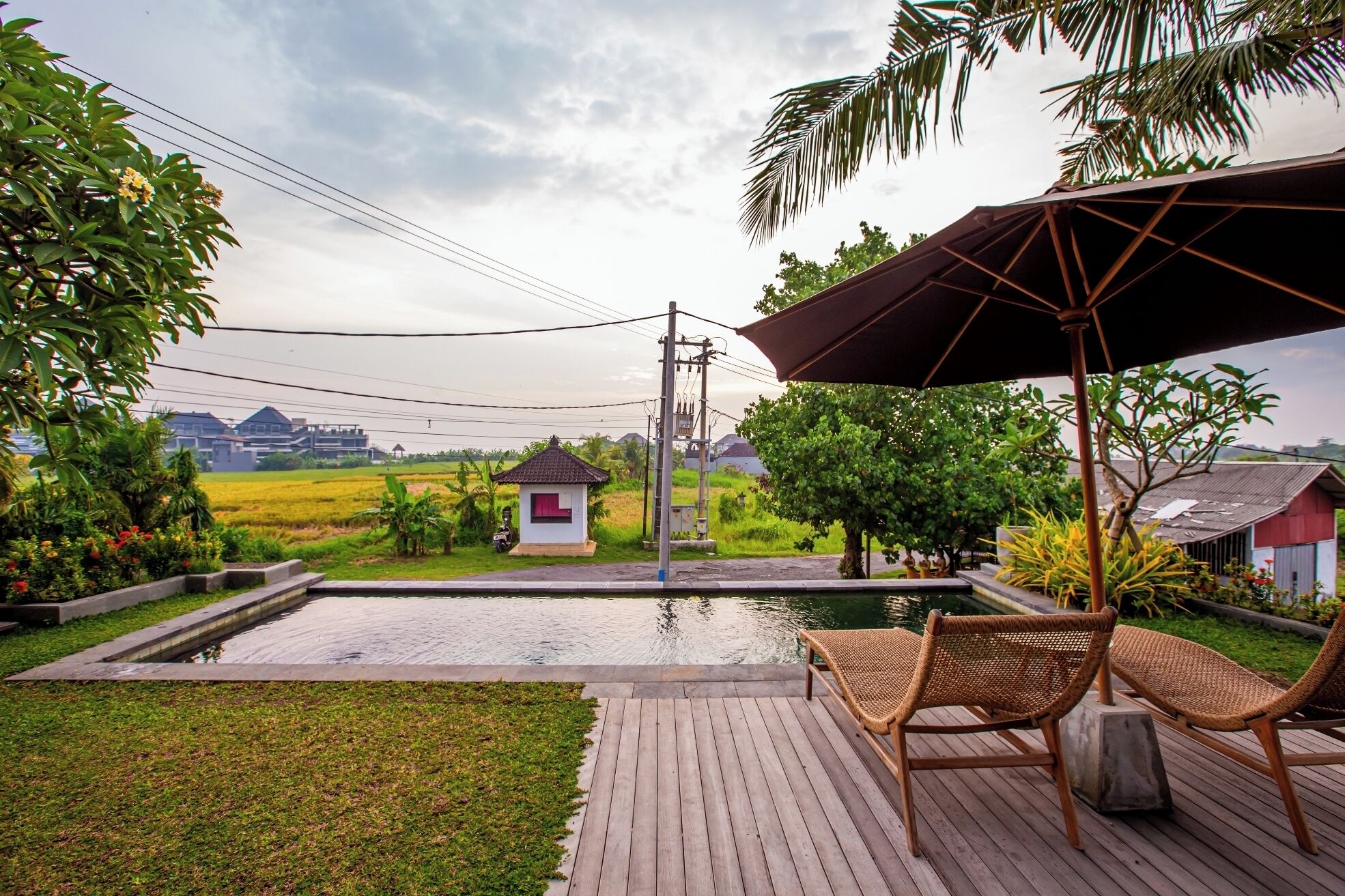 A wooden deck with two wicker chairs and a large umbrella, overlooking a small pond and a rice field.