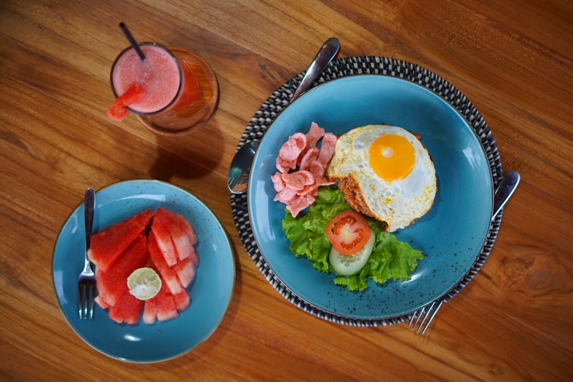 A meal with a fried egg, ham, lettuce, tomato, and watermelon on a blue plate, and a glass of a pink drink on a wooden table.