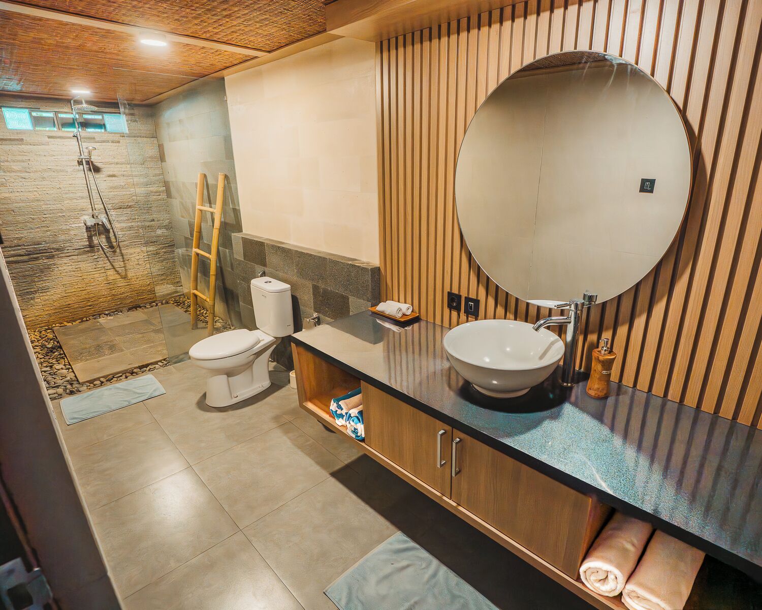 A modern bathroom with a stone shower, a toilet, a sink with a large mirror, and wooden paneling.