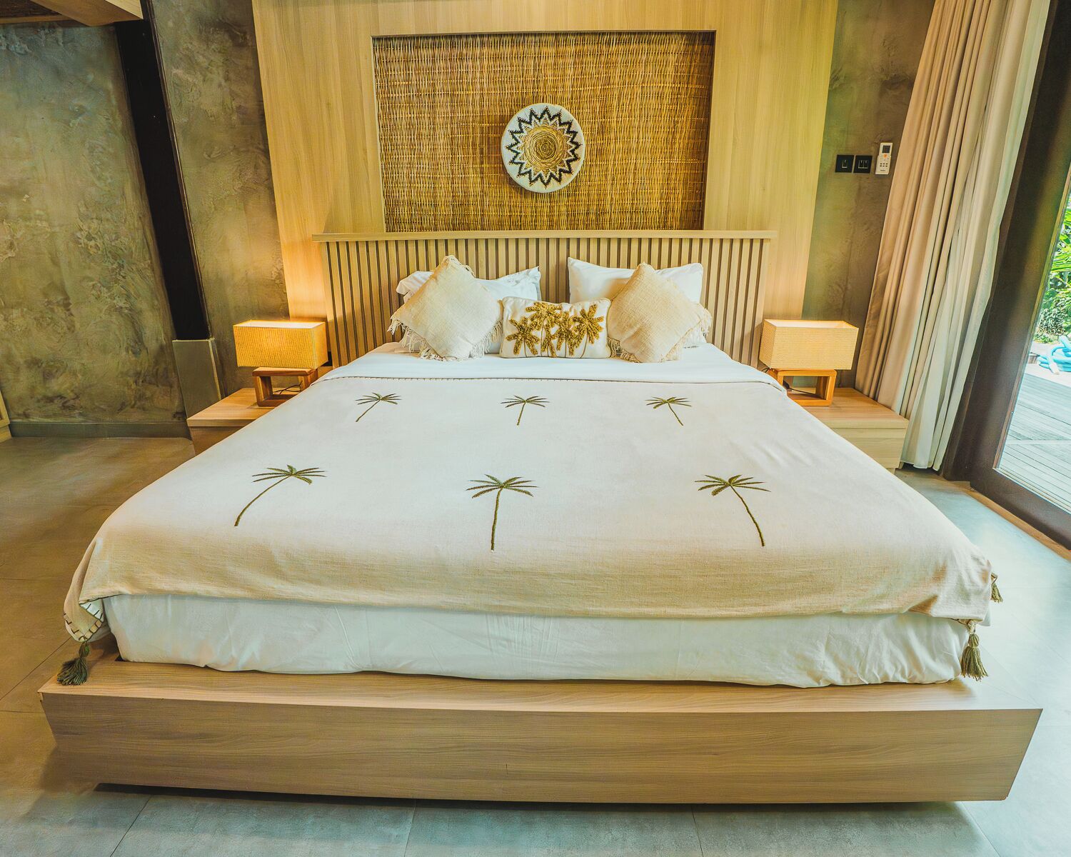 A large bed with a white bedspread and palm tree design, flanked by nightstands with lamps, a woven wall panel, and a view of greenery outside.