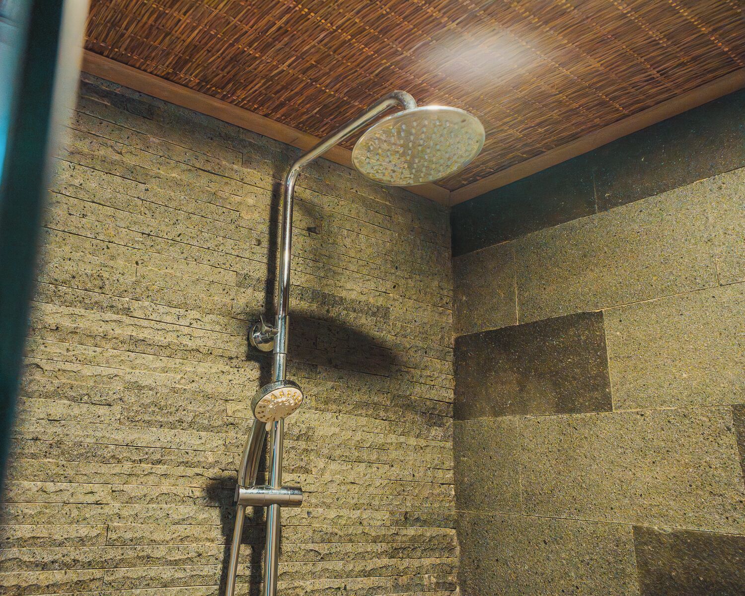 Une douche dotée d’un pommeau de douche à effet pluie, fixé sur un tuyau en métal, le tout installé devant un mur en pierre à texture marquée.