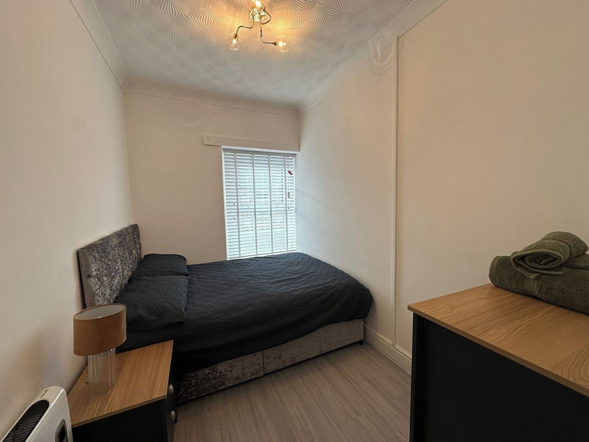 A bedroom with a bed, a bedside table, a lamp, a window with blinds, and a ceiling light fixture.