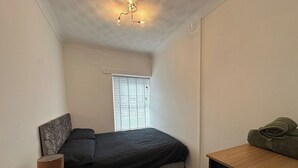 Une chambre à coucher comprenant un lit, une table de chevet, une lampe, une fenêtre avec des stores et un luminaire au plafond.