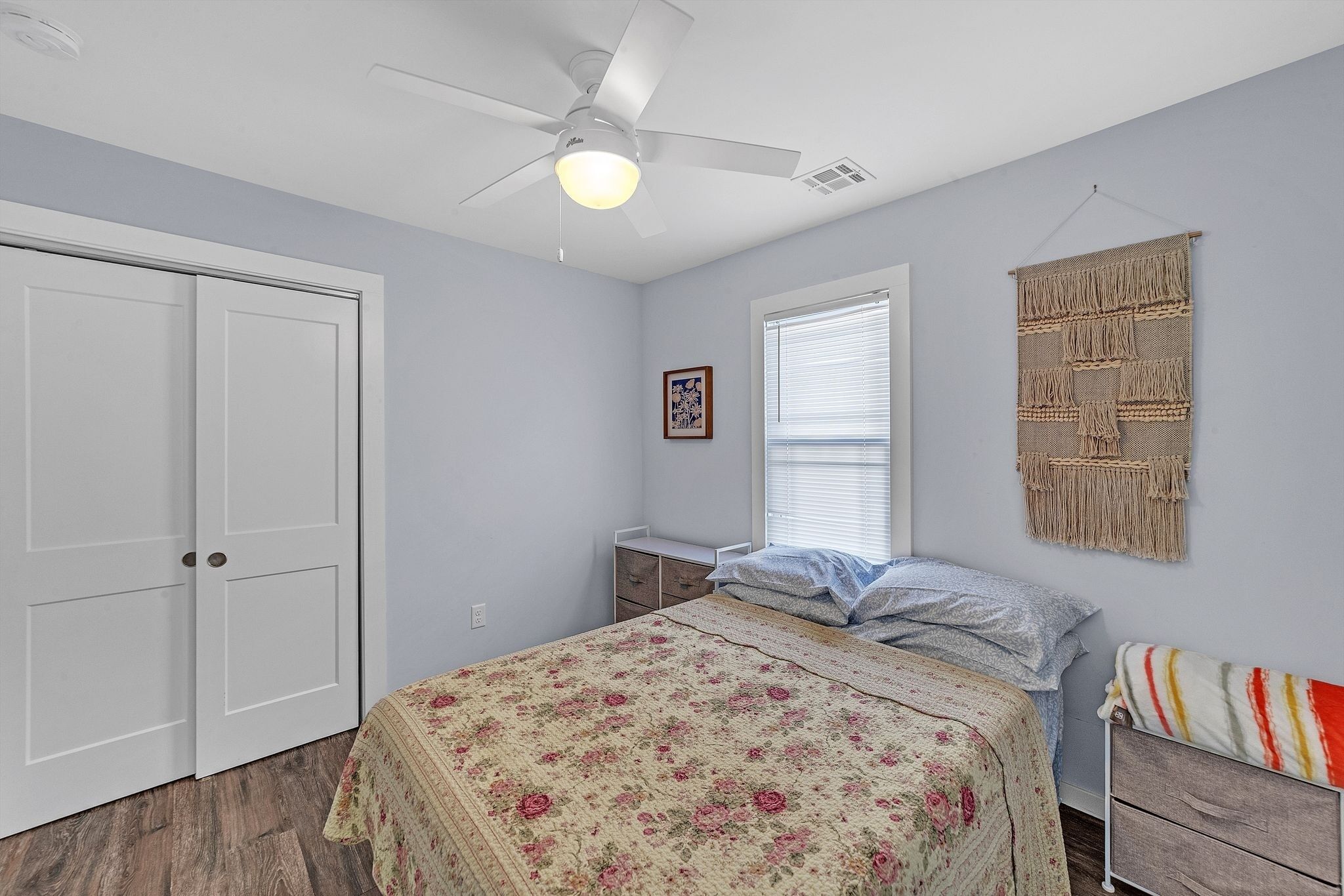 A bedroom with a bed, a ceiling fan, a window with blinds, a wall-mounted macrame piece, and a door.