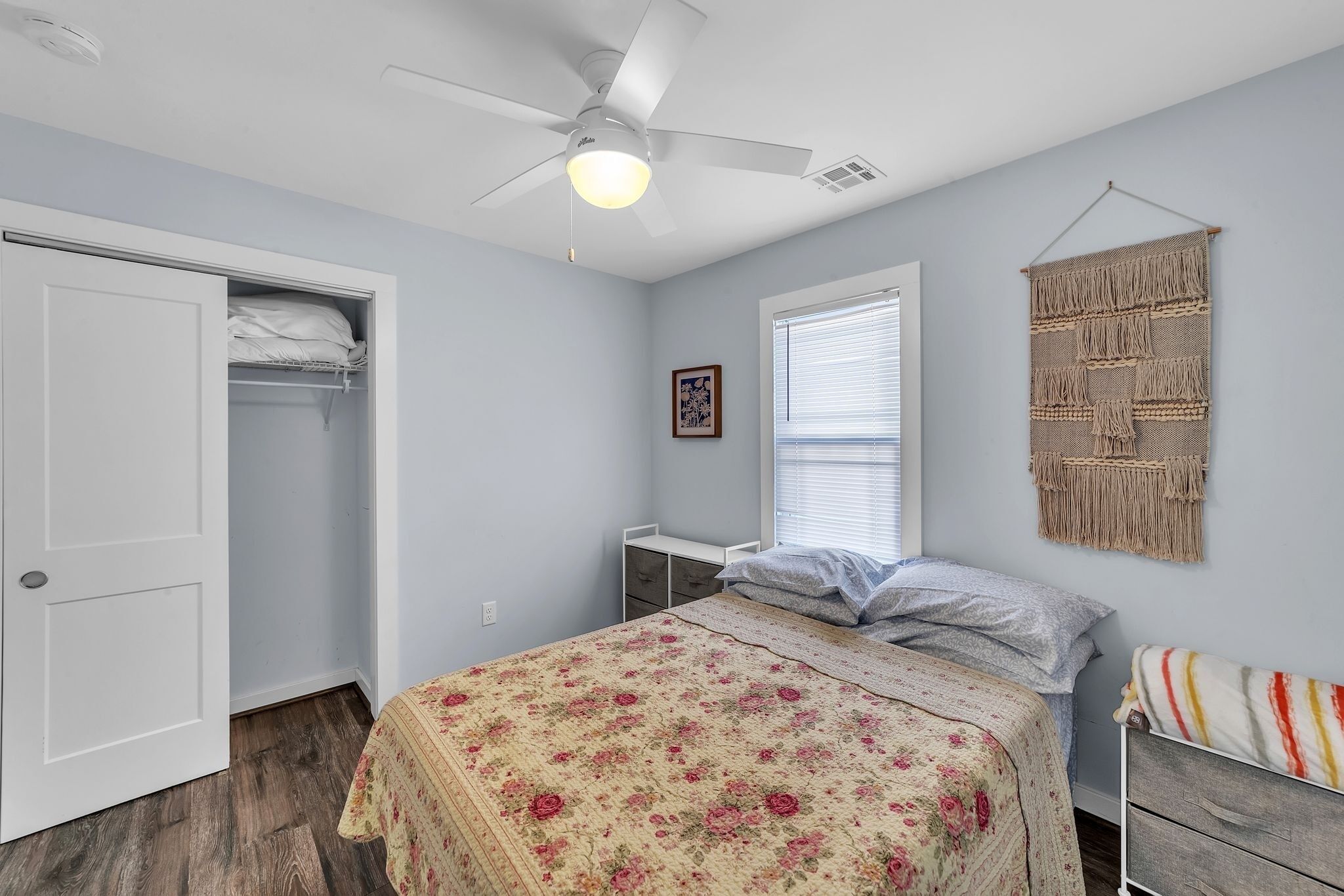 A bedroom with a bed, a ceiling fan, a wardrobe, a window, and a macrame wall hanging.