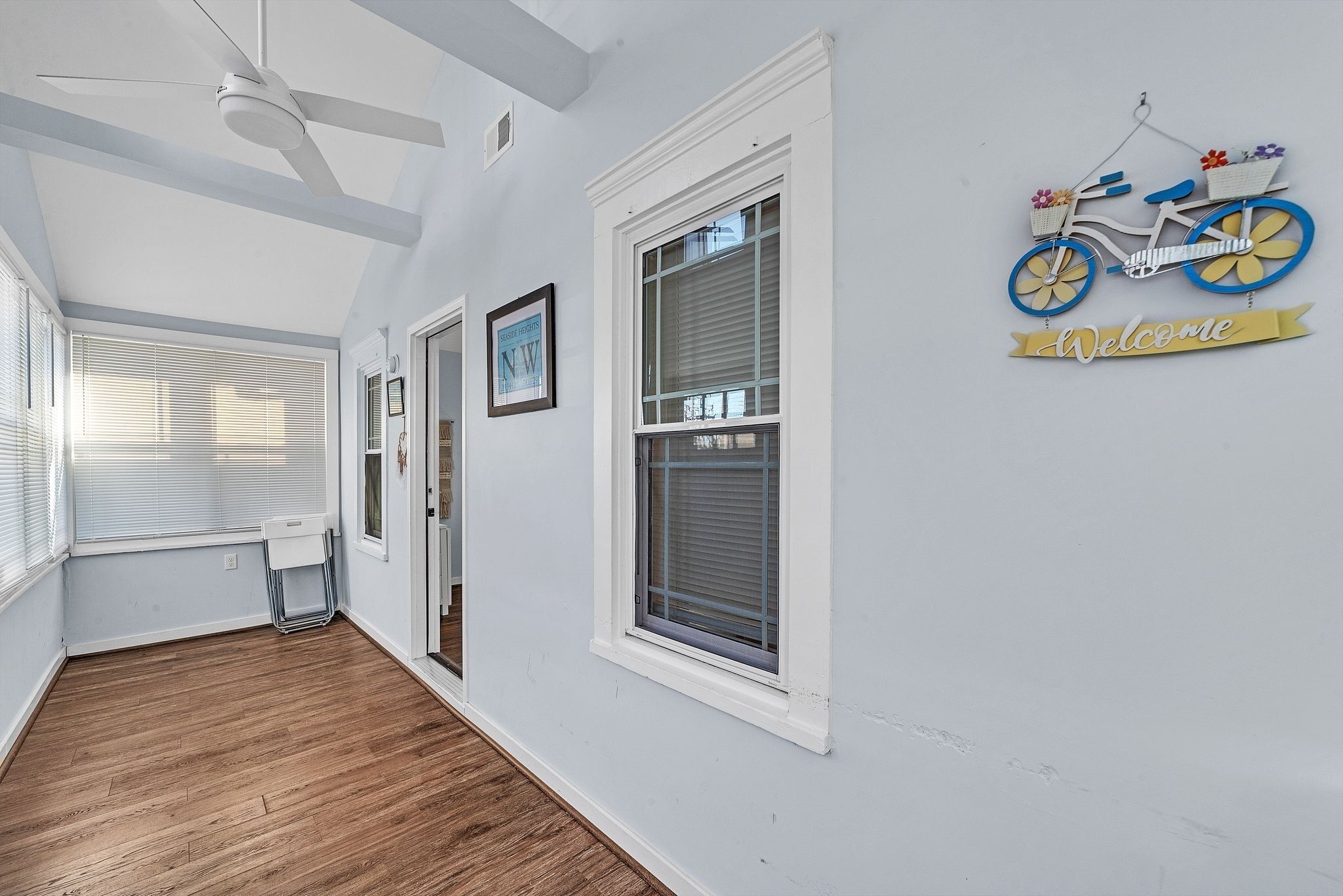 Ein Korridor mit Holzboden, weißen Wänden und einem Deckenventilator. Zwei Fenster mit Jalousien, ein gerahmtes Bild an der Wand und ein dekoratives Schild mit einem Fahrradmotiv und einem 'Welcome'-Banner.