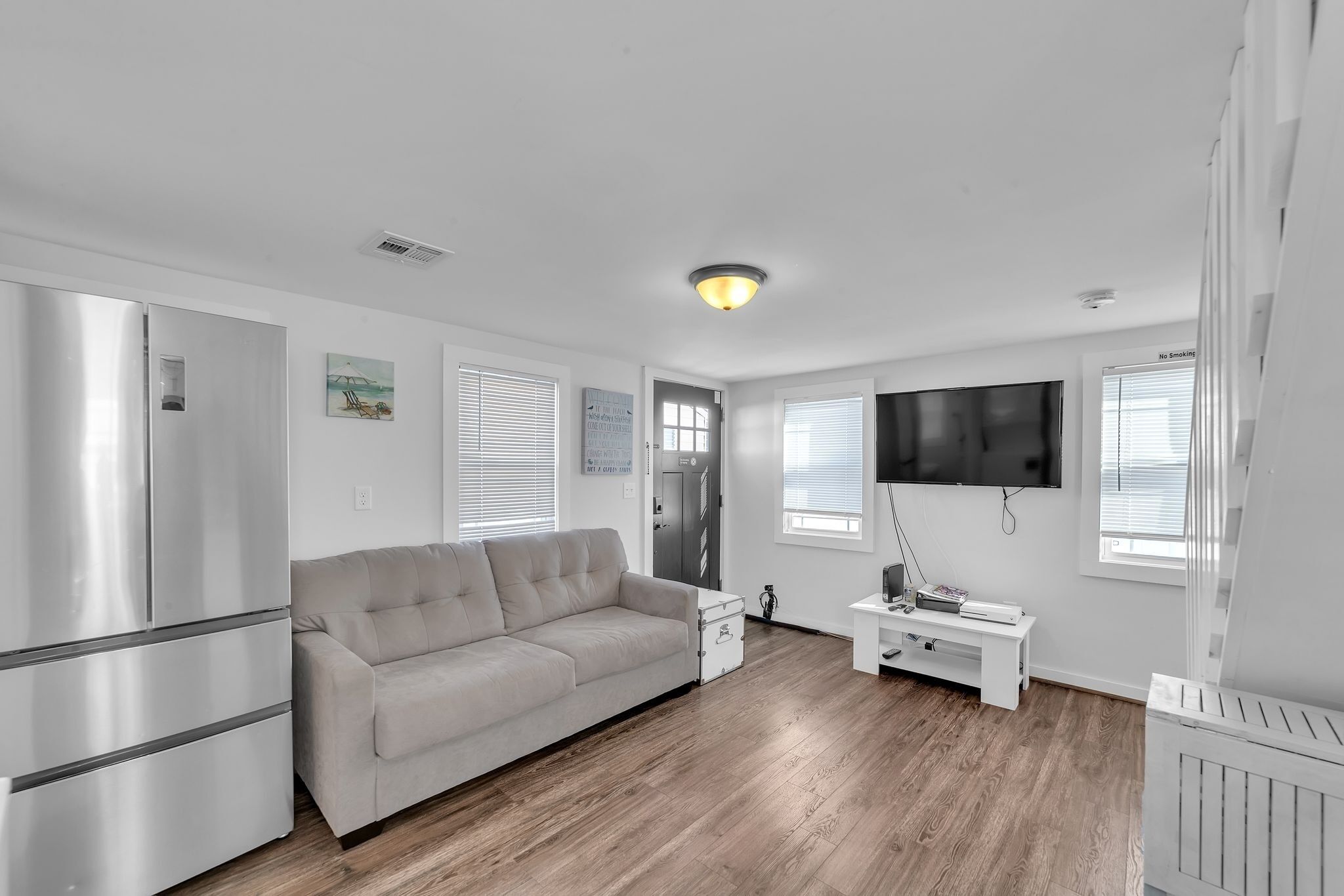 A modern, well-lit living room with a beige sofa, a flat-screen TV mounted on the wall, and a white cabinet.