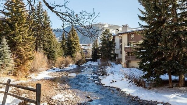 Une rivière traverse un paysage enneigé, avec des bâtiments d’un côté et une montagne en arrière-plan.