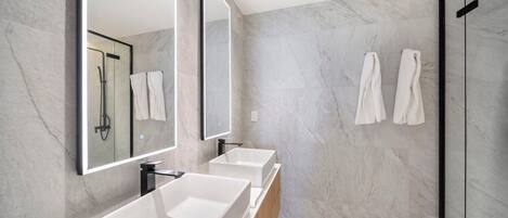 Un baño moderno con dos lavamanos blancos, un espejo grande y una cabina de ducha con mampara de cristal.
