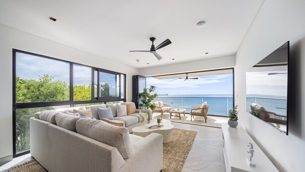 Un salon moderne avec un canapé, une table basse et des fauteuils, de grandes fenêtres offrant une vue sur la mer et un ventilateur de plafond.