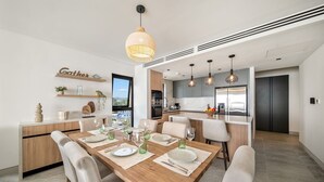 A modern kitchen with a central island, a dining area with a wooden table, and a view through a window. - Allure 505 3 Bedroom Apts (Bridgetown)