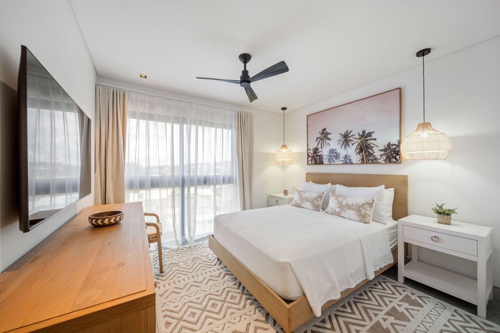 A hotel room with a bed, bedside tables, a desk, a chair, a ceiling fan, a large window with curtains, and a wall-mounted artwork.