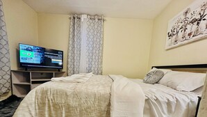 A neatly made bed with a white comforter and pillows, a television on a stand, a patterned curtain, and a wall art piece. - Charming 2-bedroom house in Aberdeen with WiFi and AC (Aberdeen)