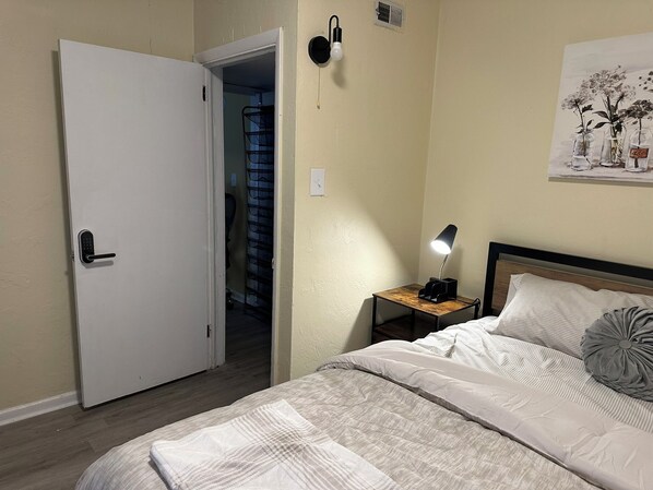 Una camera da letto con un letto, un comodino con una lampada, una porta e un punto luce a parete.