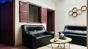 A living room with two black leather sofas, a coffee table, and a potted plant. There are two windows with shutters, a skylight, and a wall-mounted hexagonal shelf. - Apartment in the center of Tiznit (tiznit neuf)