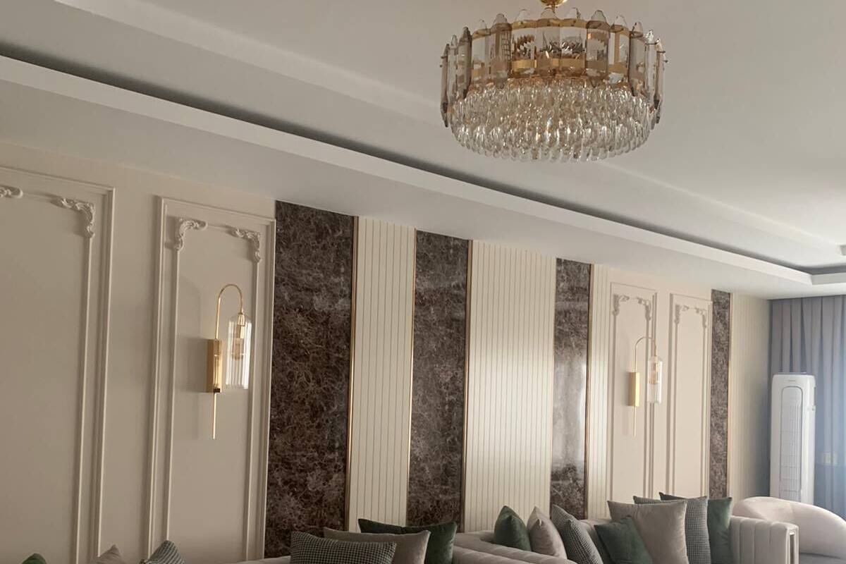 A spacious living room with a long, upholstered sofa, a decorative chandelier, and a marble accent wall.