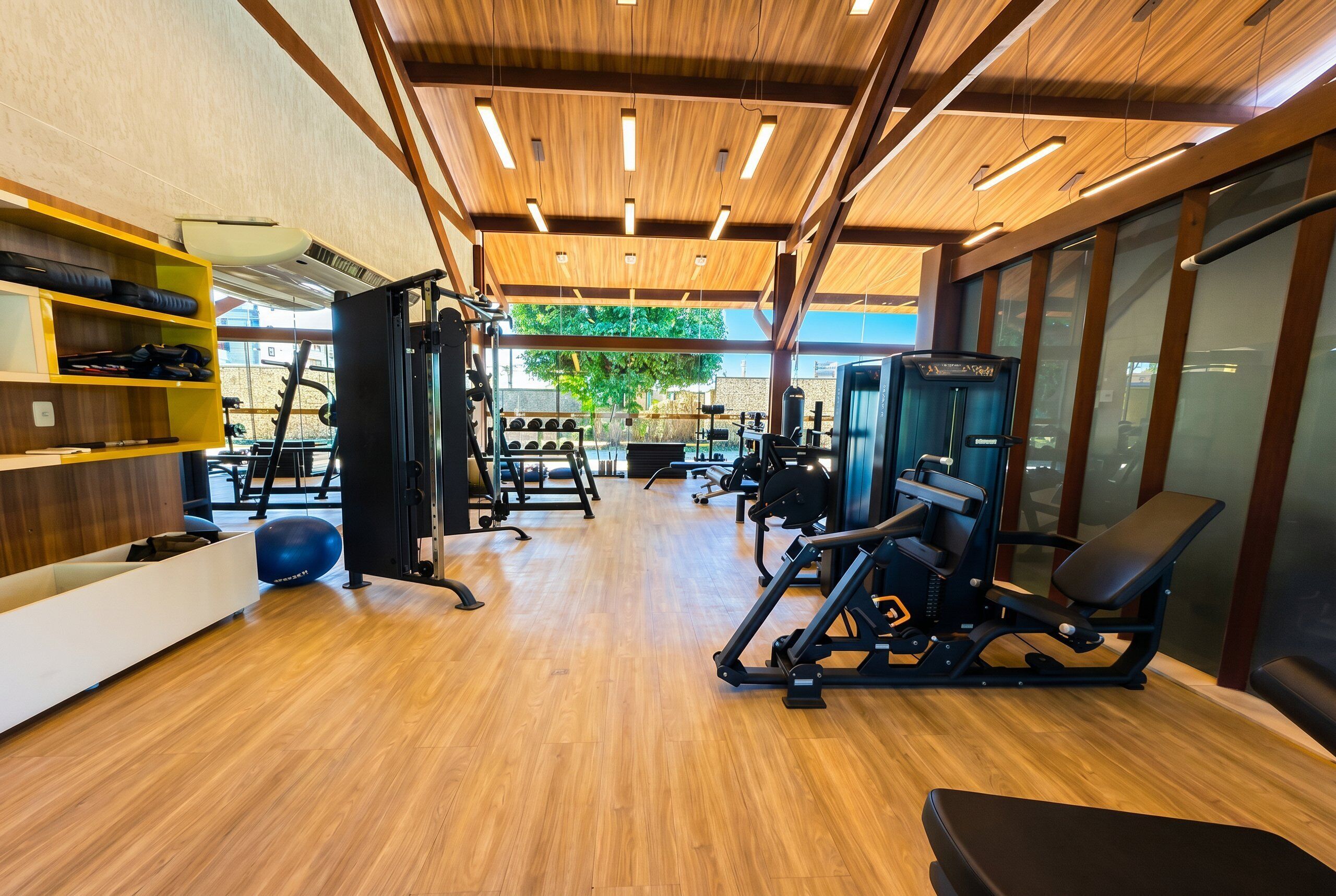 Academia com piso de madeira, equipamentos de exercício e vista para o exterior.