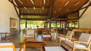 An indoor area with wooden tables and chairs, a pool table, and large windows allowing natural light. - Porto de Galinhas, Malawi Beach Resort - Flat SKY (Ipojuca)