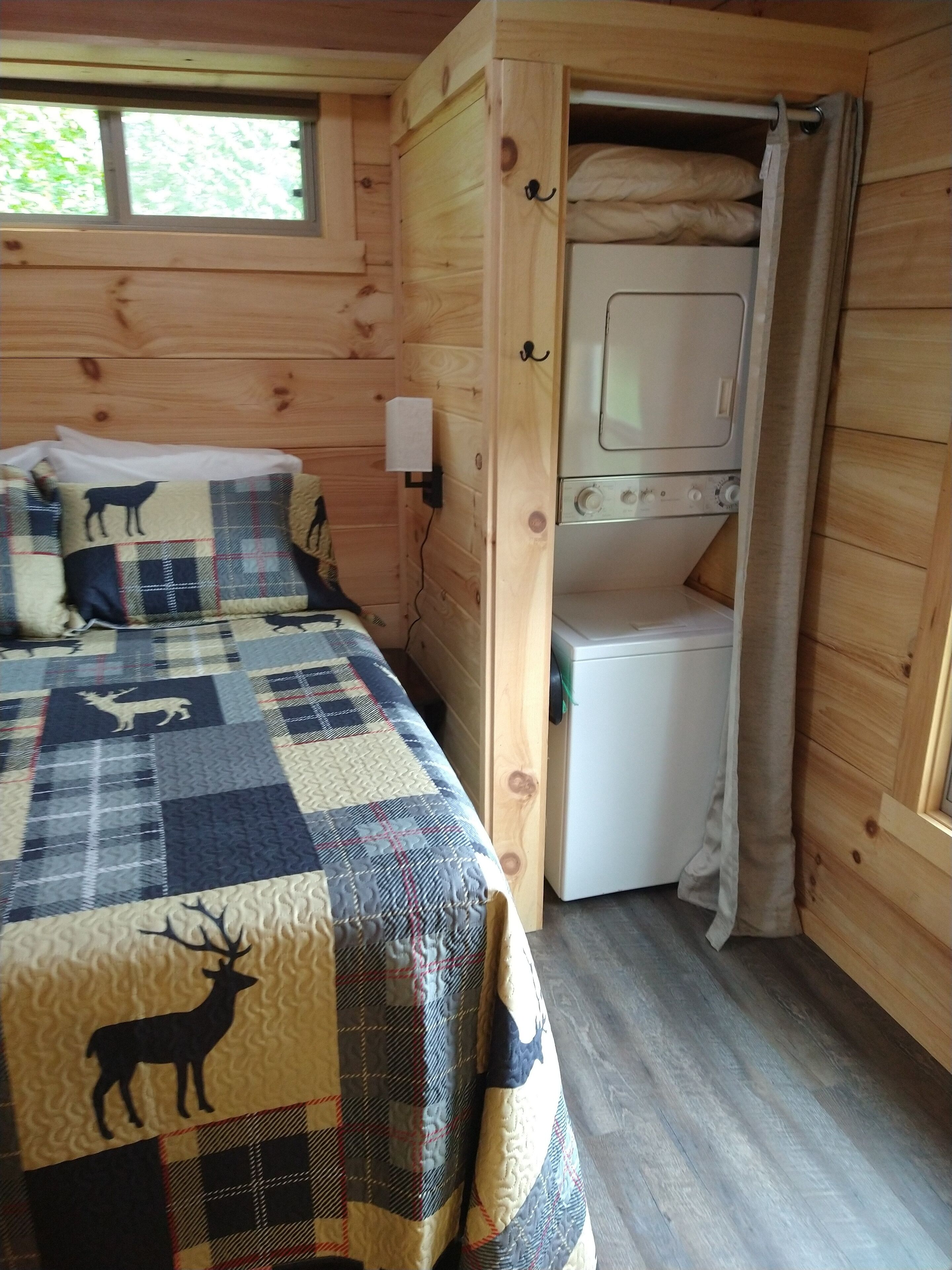 Habitación de cabaña de madera con una cama que tiene un colcha estampada con animales, una lavadora blanca pequeña y una ventana con vista a árboles.