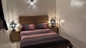 Un dormitorio con cama de madera, ropa de cama estampada, dos lámparas fijadas a la pared y suelo de baldosas de mármol.