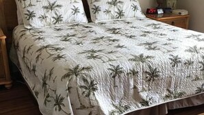 Ein Bett mit tropischem Palmblattmuster, ein hölzernes Kopfende, ein Nachttisch mit Lampe und eine digitale Uhr.