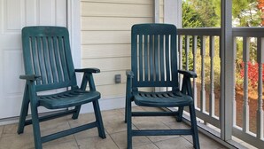 Zwei grüne Adirondack-Stühle auf einer gefliesten Veranda mit einer weißen Tür und Blick auf Bäume und Sträucher.