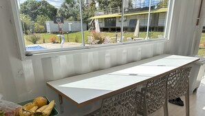 A room with a long table, chairs, a window with a view of a basketball court, and a green bin with bananas and onions. - Villela House at Villela Home Stay (Lago Sul)