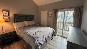 Quarto com cama, mesa de cabeceira, cômoda e janela com cortinas.