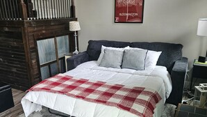 Um quarto com uma cama, um sofá cinzento, um cobertor xadrez vermelho e branco, um tapete cinza, uma escada de madeira e um mapa fixado na parede.